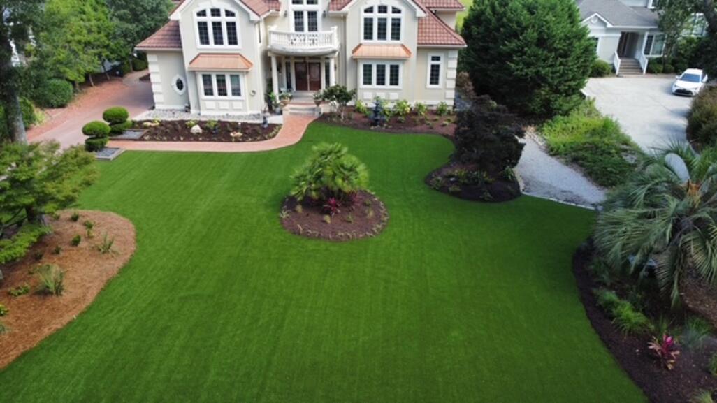 Aerial view of a luxury coastal home with a lush synthetic turf front lawn and manicured planting beds