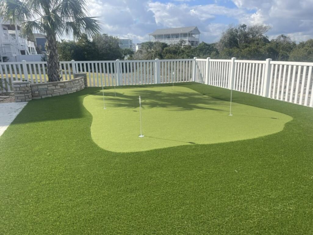 Backyard synthetic turf putting green framed by a white fence and coastal palms
